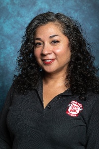 Picture of Britney Stout,Administrative Assistant, Intermediate, American Indian Program, Asian and Pacific Islander Programs and LGBT+ Programs.  The image is a professional portrait of a person with curly, shoulder-length hair. They are smiling gently and looking slightly to the right. The background of the image is a textured blue backdrop, giving a studio photo appearance. The person is wearing a black collared shirt that features an embroidered red and white logo on the left side. The logo has the text "NMSU" in the center, with additional text around it.